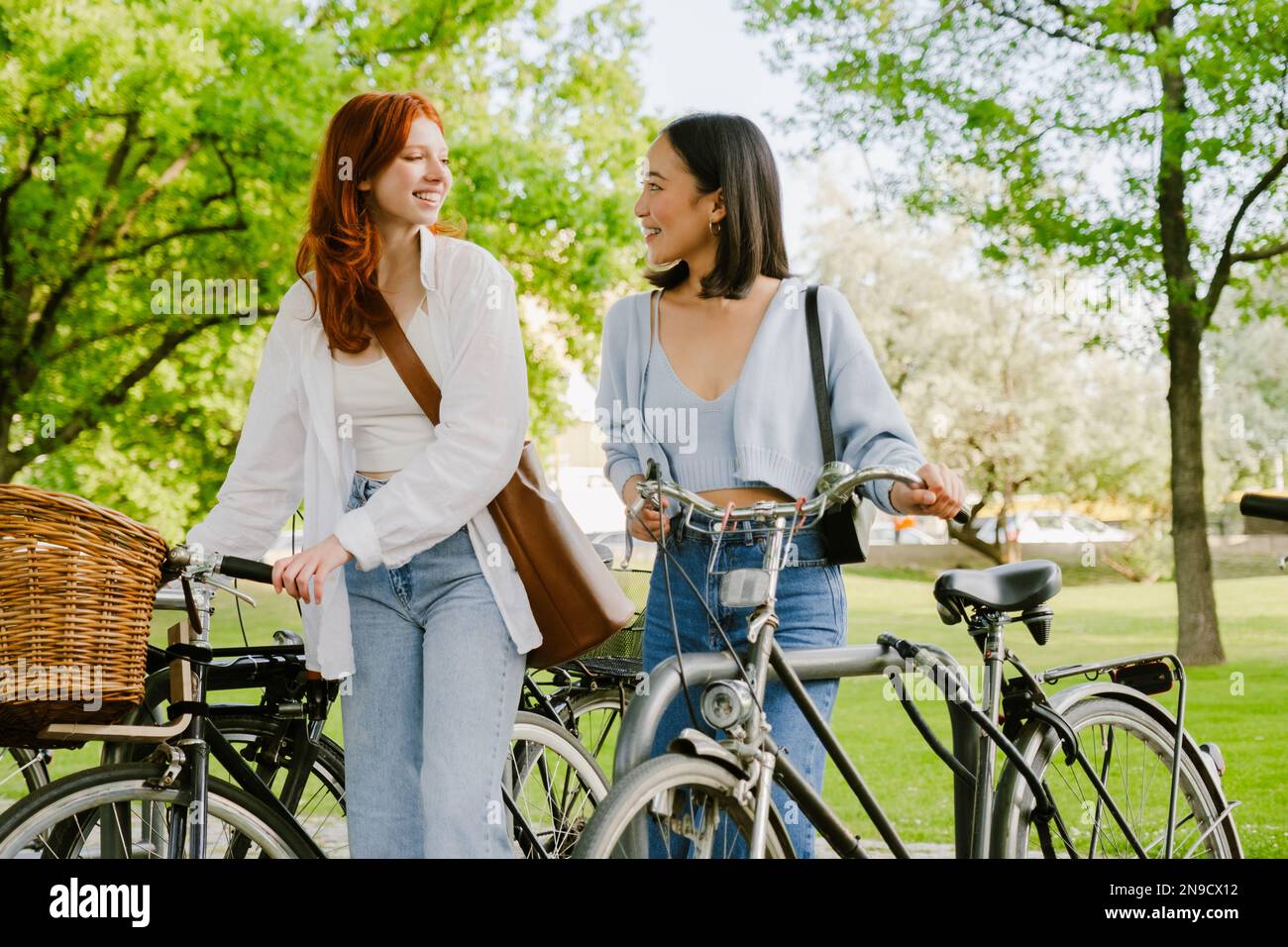Two young beautiful smiling happy girls walking with bicycles in park and looking on each other Stock Photo