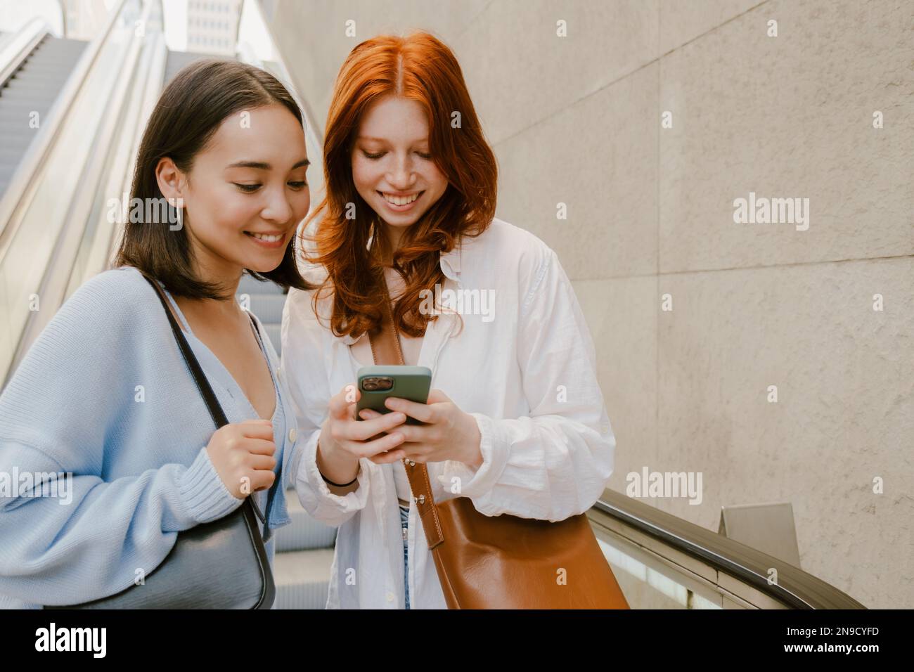 Young multinational women smiling and using cellphone while standing indoors Stock Photo