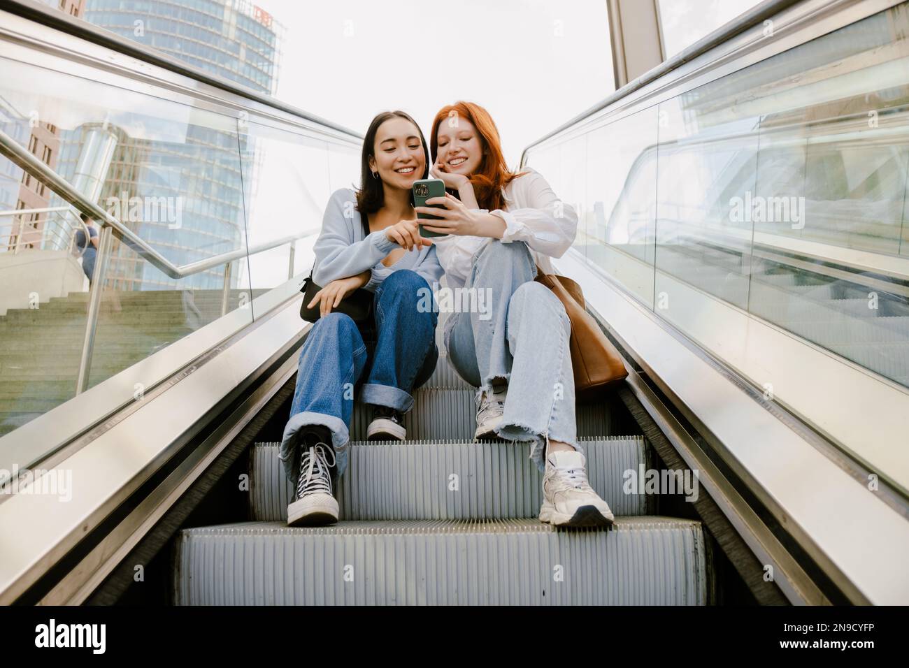 Young multinational women smiling and using cellphone while sitting on escalator Stock Photo
