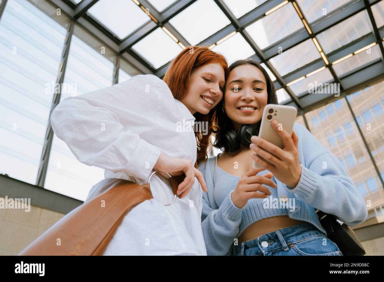 Young multinational women smiling and using cellphone while standing indoors Stock Photo