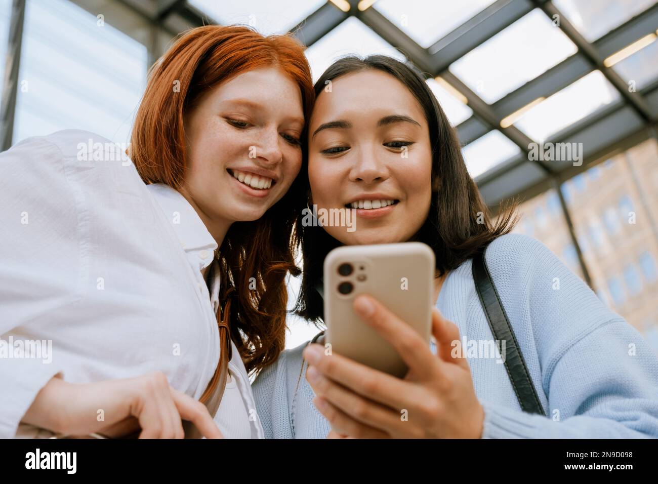Young multinational women smiling and using cellphone while standing indoors Stock Photo