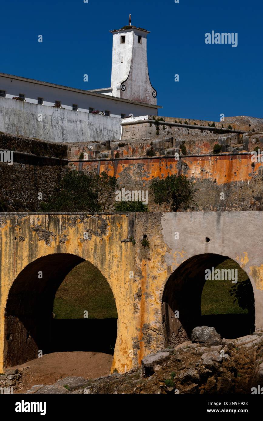 Yellow, orange and white: the moat bridge, battered ramparts and Torre de Vigia watchtower, decorated with spiral scrolls, above one of the blocks of prison cells built in the 1950s inside the mid-1500s Fortaleza or Fortress at Peniche, on central Portugal’s Atlantic coast.  Political opponents of the 1933-74 Salazar government, including communist leader Álvaro Cunhal, were held here, often in high security isolation cells. Stock Photo
