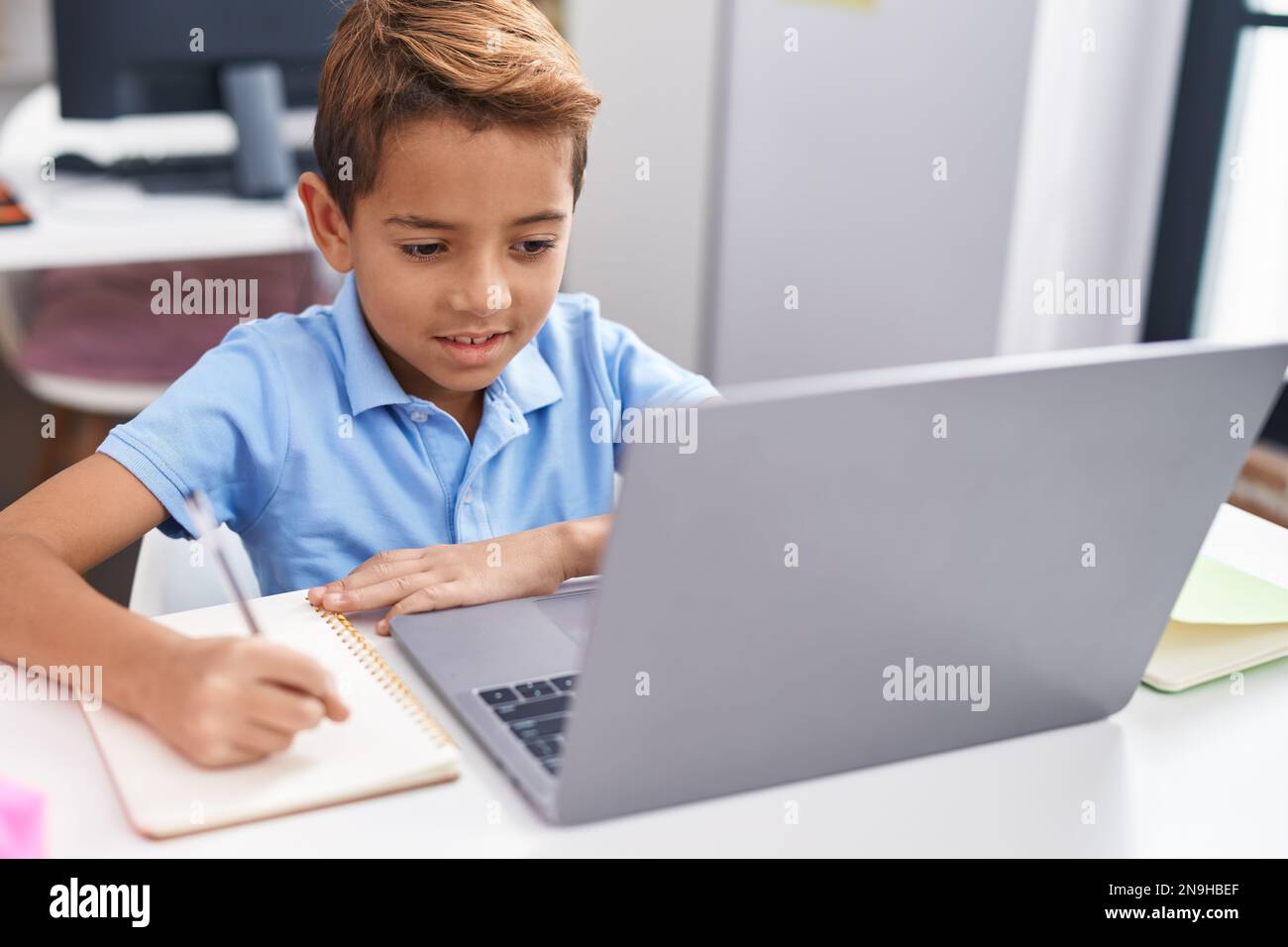 Adorable hispanic boy student using computer writing on notebook at ...