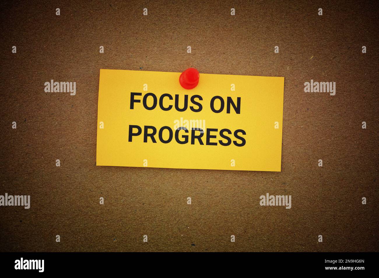 A yellow paper note with the phrase Focus On Progress on it pinned to a cardboard background. Close up. Stock Photo