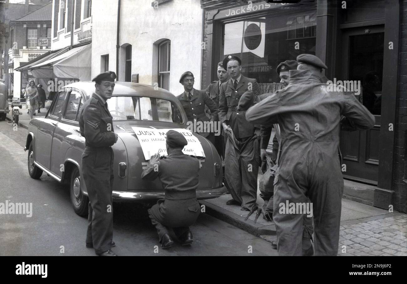 1950s, historical, a number of army officers gathered on a pavement, with one sticking on the back boot of a car of the era parked in a town high street, a large Learner driver notice, saying a 'Just Married'. The fun wedding car notice reads; 'No Hand Signals', 'Driver in Love', Stock Photo