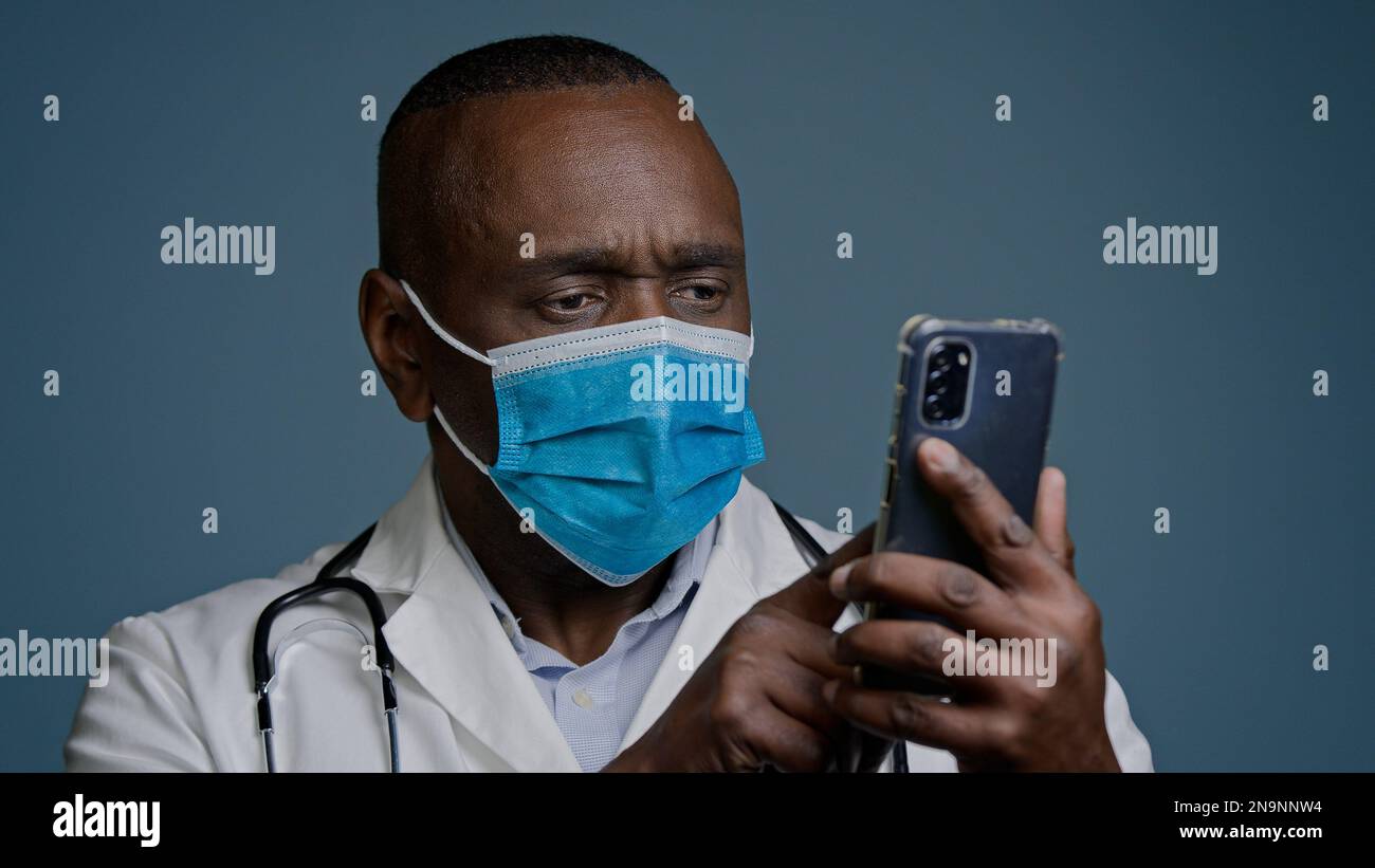 Close-up experienced mature doctor in protective mask and white medical gown reads message on mobile phone checks information on Internet concentrated Stock Photo