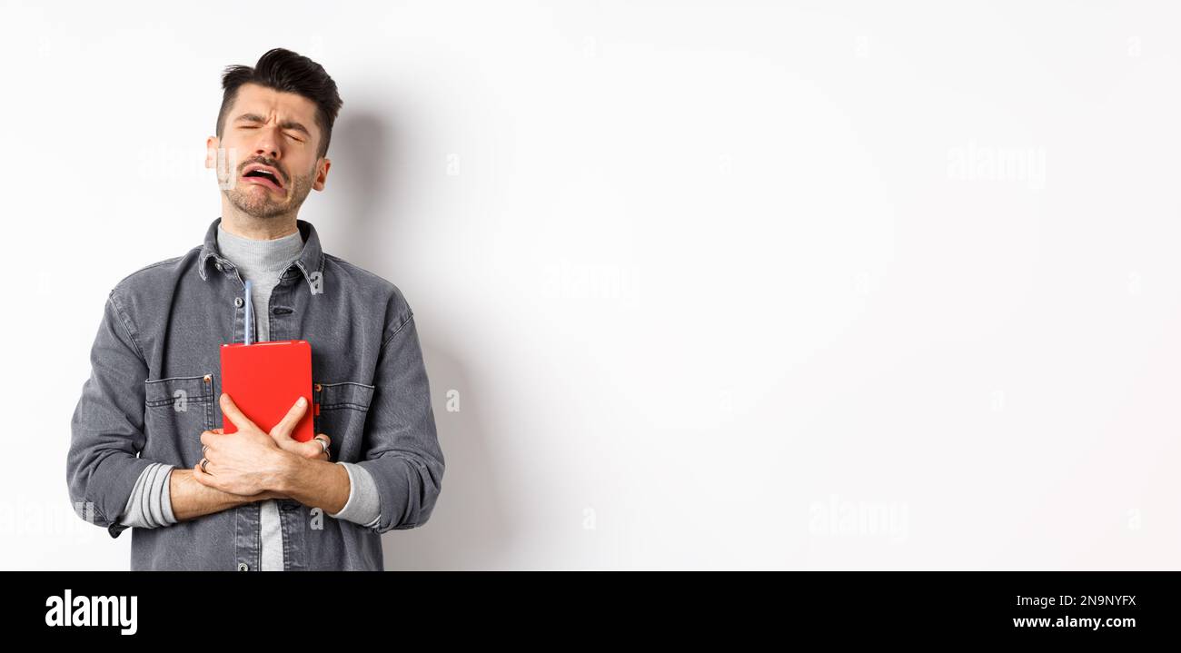 Sad cryig man holding red diary and sobbing, miserable guy carry journal with him, standing against white background Stock Photo