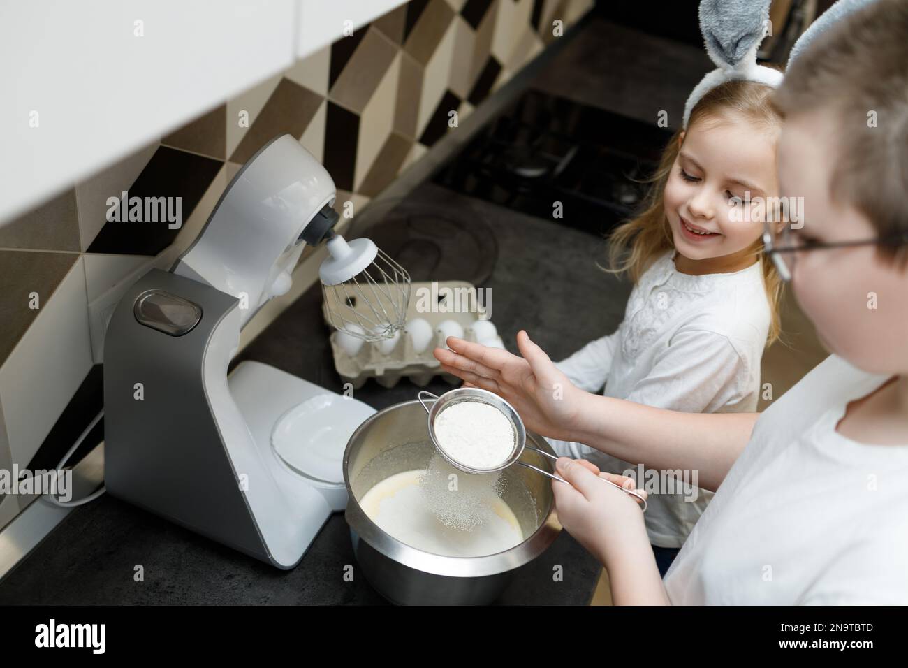 Brother sifting flour, preparing dough for cookies, cross buns. Sister in bunny ears intrested looking at process. Easter, family support concept Stock Photo