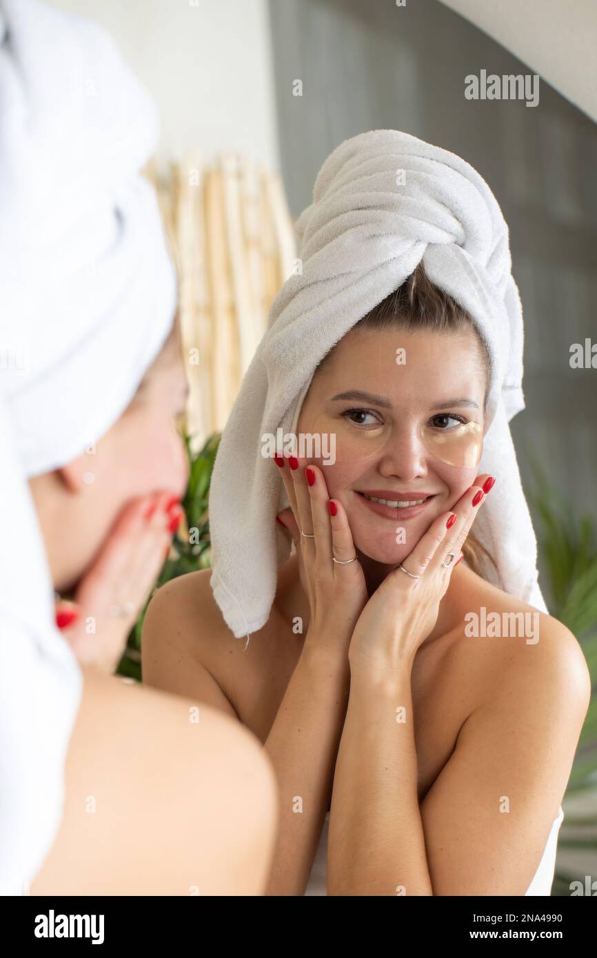 Attractive woman with patches under her eyes looks at her reflection in the mirror while smiling.  Stock Photo