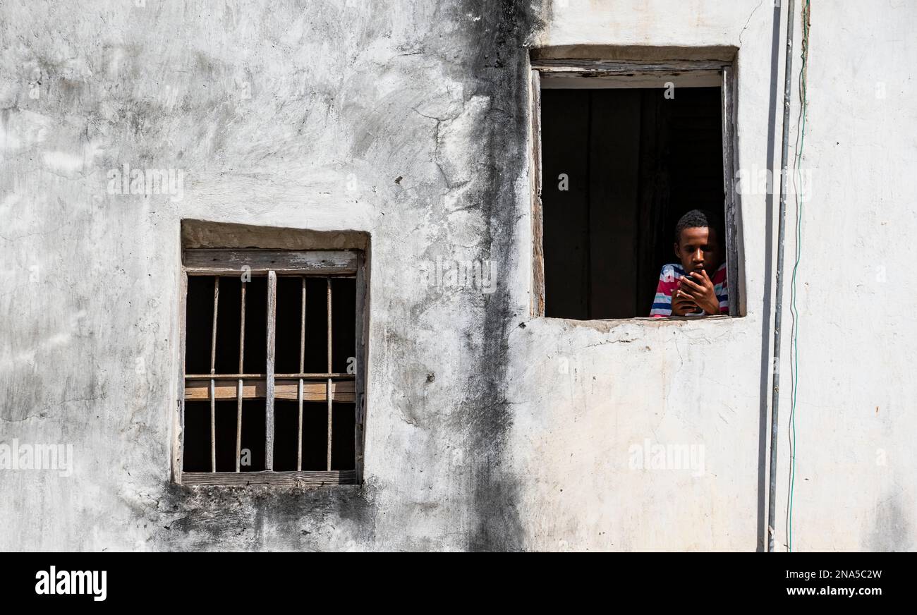 Boy in window holding smart phone, Stone Town of Zanzibar; Zanzibar City, Unguja Island, Zanzibar, Tanzania Stock Photo