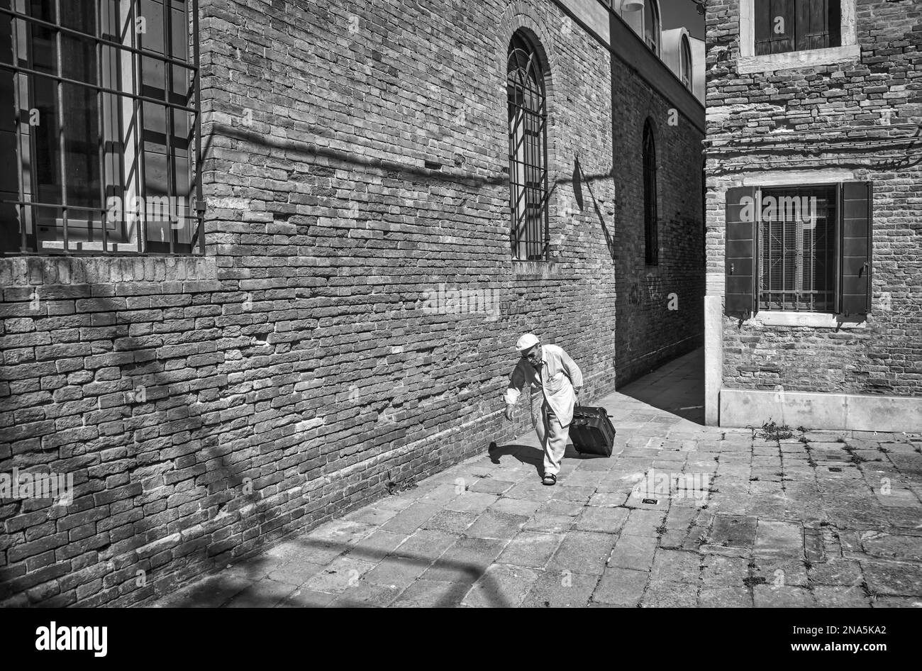 Man rolls a suitcase beside the brick wall of a building; Venice, Italy Stock Photo