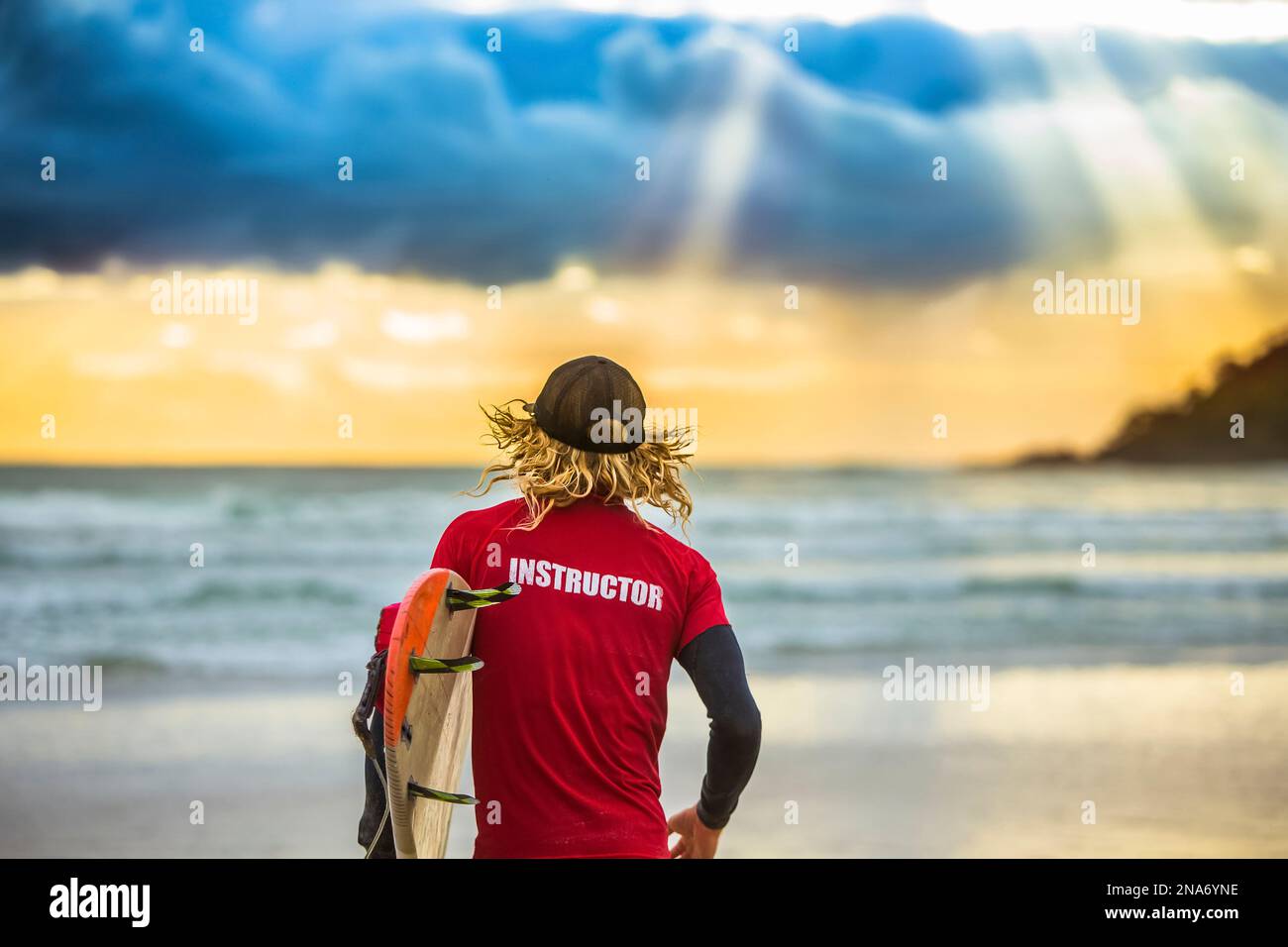 View taken from behind of a surfing instructor looking out at ocean greeted by a beautiful sunrise as he prepares to set out for an early morning surf Stock Photo