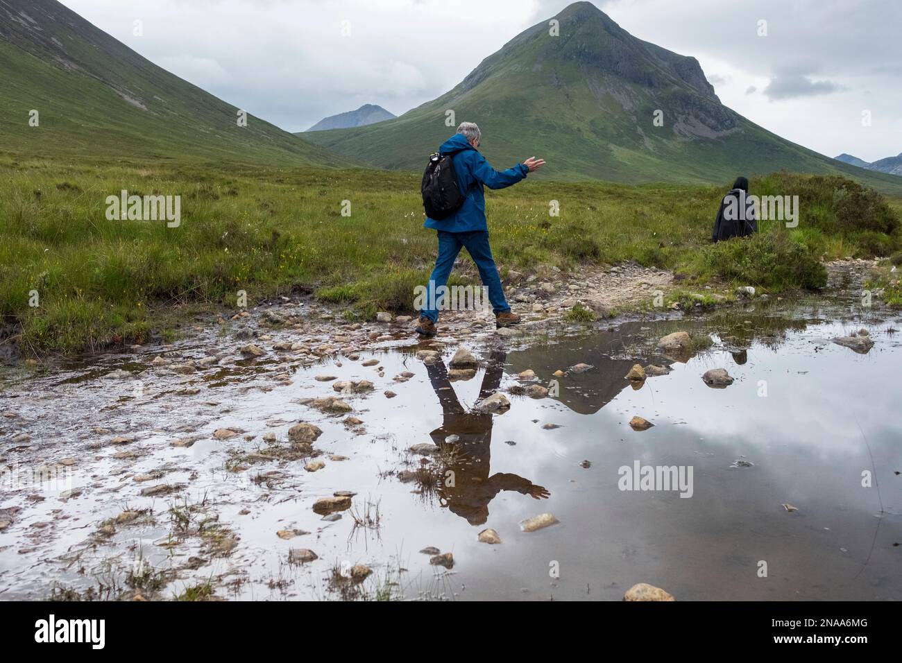 A hiker traverses a small stream along a trail in the Cuillin Mountains, Sligachan, Isle of Skye, Scotland. Stock Photo