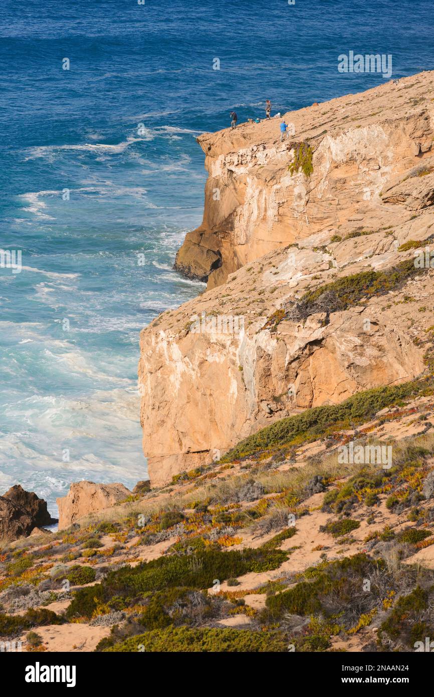 Tourists stand on a sloping rugged ridge overlooking cliffs and coastline of the Atlantic Ocean, Praia de Monte Clerigo and Southwest Alentejo and ... Stock Photo