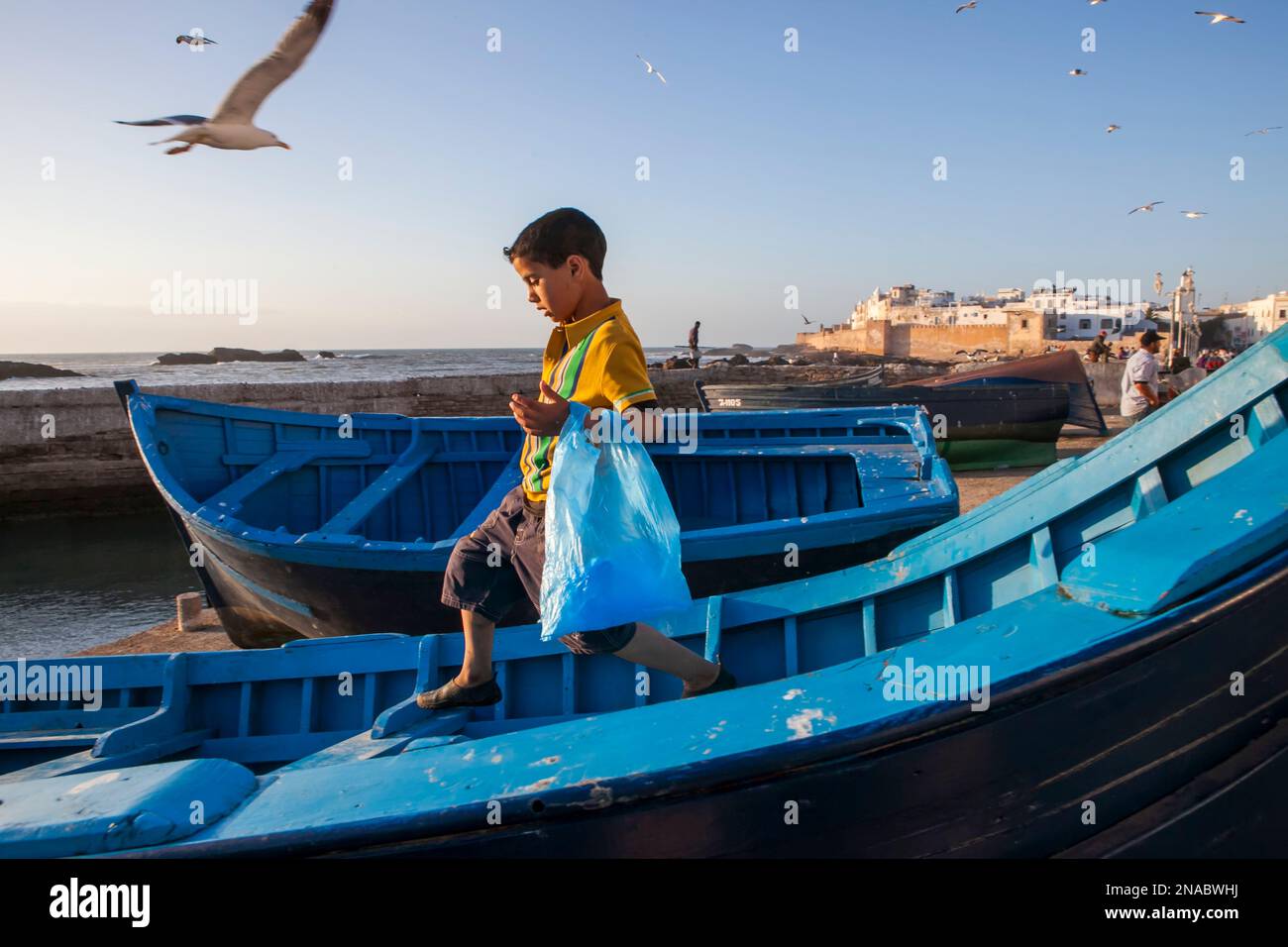 A young boy gingerly traverses a boat on the waterfront of Essaouira, Morocco; Essaouira, Morocco Stock Photo