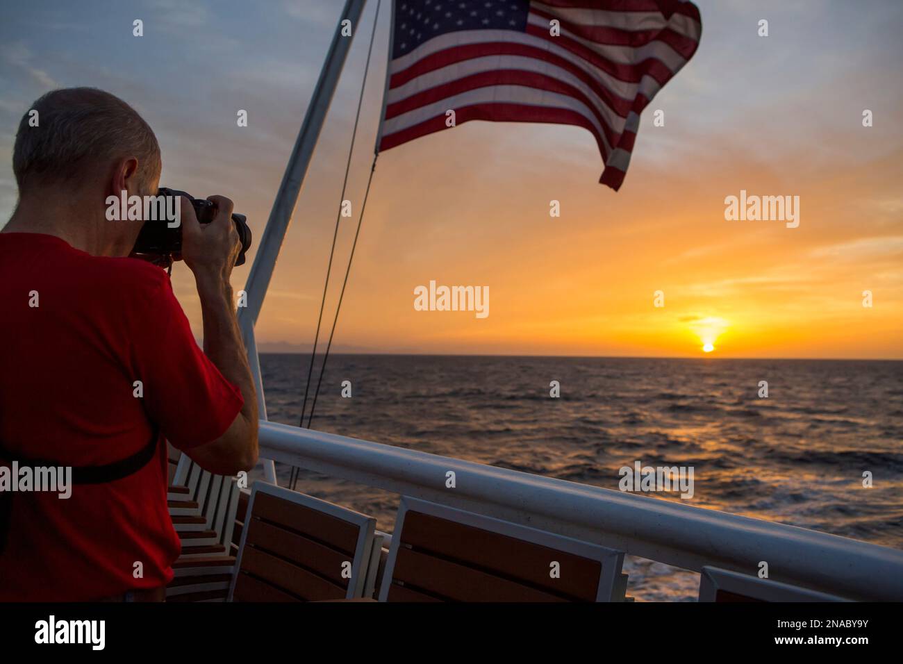 Amidst a colorful morning sky near Panama, a passenger aboard an expedition ship photographs the sunrise as an American flag billows in the wind Stock Photo