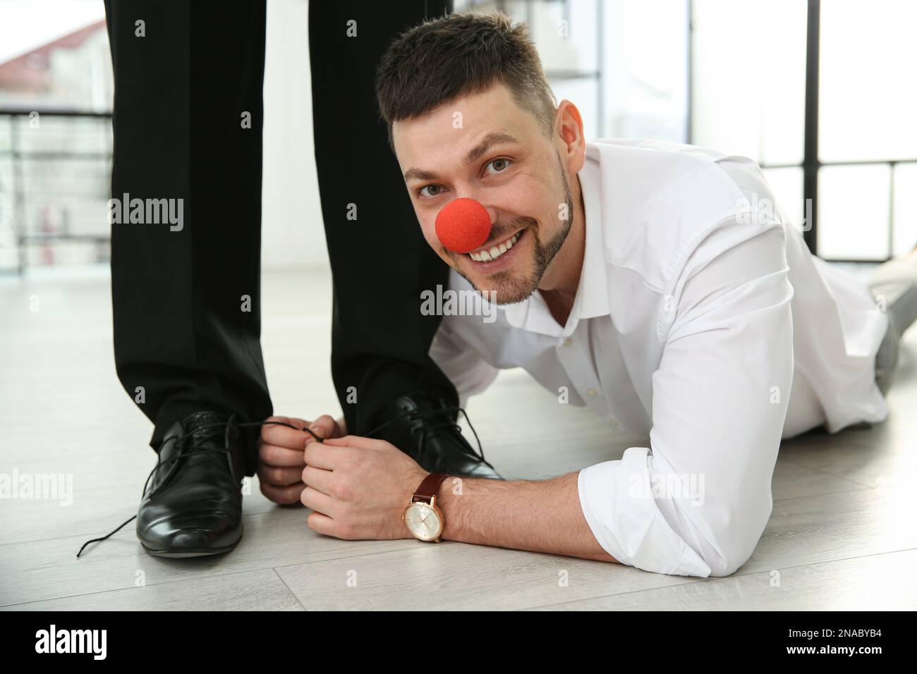 Man with clown nose tying shoe laces of his colleague together in