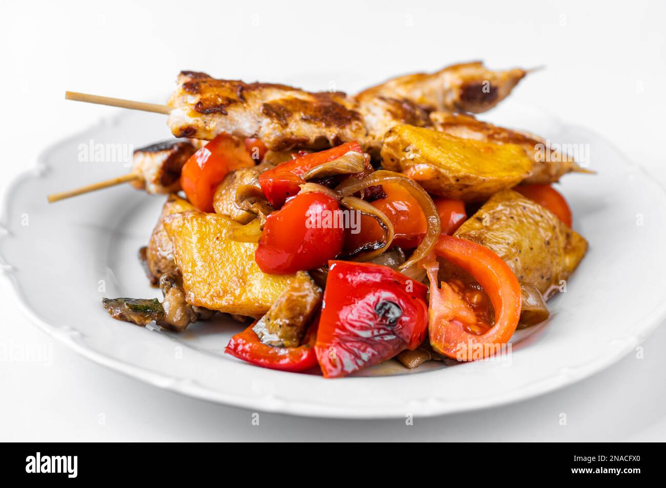 Souvlaki, pork meat on skewer on baked juicy vegetable on white plate on white background. Delicious greek lunch or dinner, closeup. Stock Photo