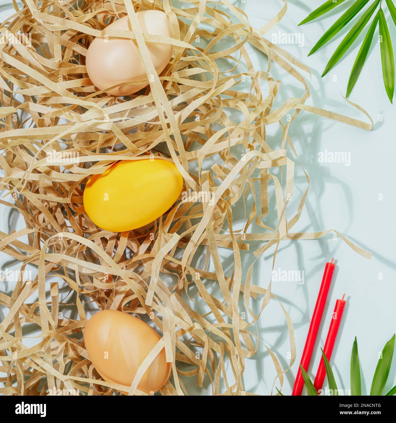 Easter holiday composition with colored eggs on shredded paper, red church candles and palm leaf on blue background. Springtime. Congratulatory easter Stock Photo