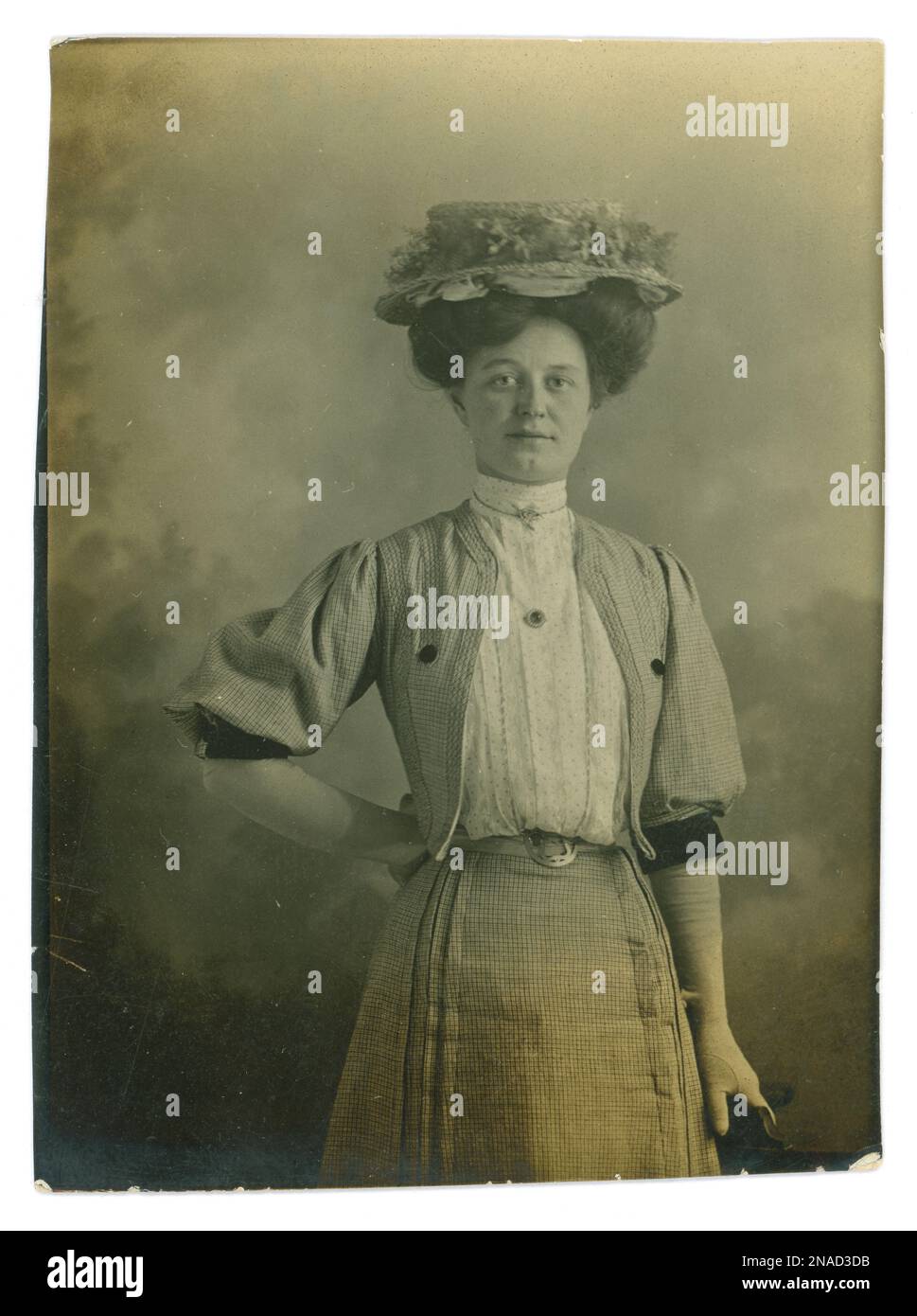Original Edwardian photo of a pretty working class lady called Amy. She may have been a lady's maid. She is wearing a straw hat, big hairstyle, wishbone style brooch, white gloves,  belt around a very slime waist, fine pleated cotton summer blouse with small dots, with a high neckline, bolero type jacket with full sleeves to elbow and matching skirt. Dated 5 June 1907. Stock Photo