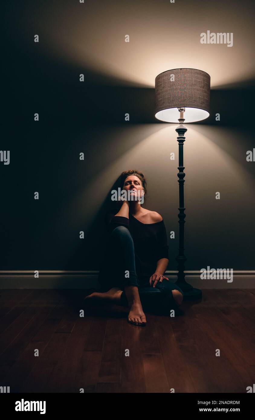 Pretty woman sitting alone on the floor beside a lamp in dark room ...