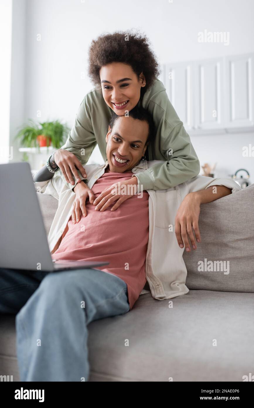 African american woman hugging tattooed boyfriend near laptop on couch ...