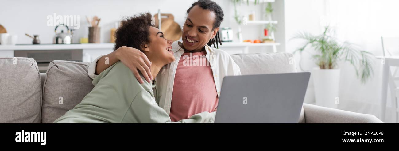Smiling african american woman hugging boyfriend near laptop on couch ...
