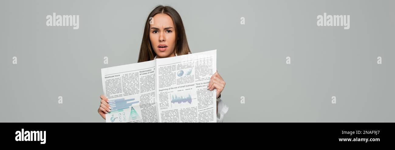 shocked young woman holding newspaper while looking at camera isolated on grey, banner,stock image Stock Photo