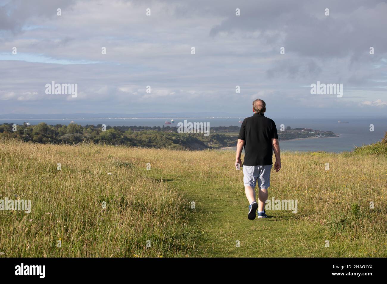 A man walks along the top of a hill on a grass path. He looks out over the sea and land in the distance. It looks relaxing and inviting for a hike. Stock Photo