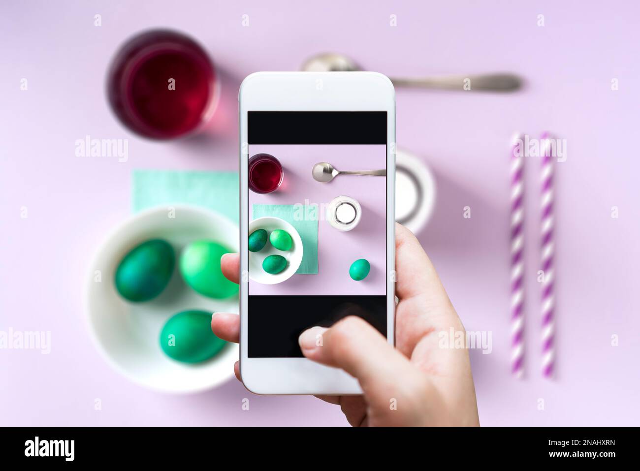 Hand taking photo with smart phone. Green dyed easter eggs, spoon, milk, straw and food coloring  on table top flat lay. Purple background. Stock Photo
