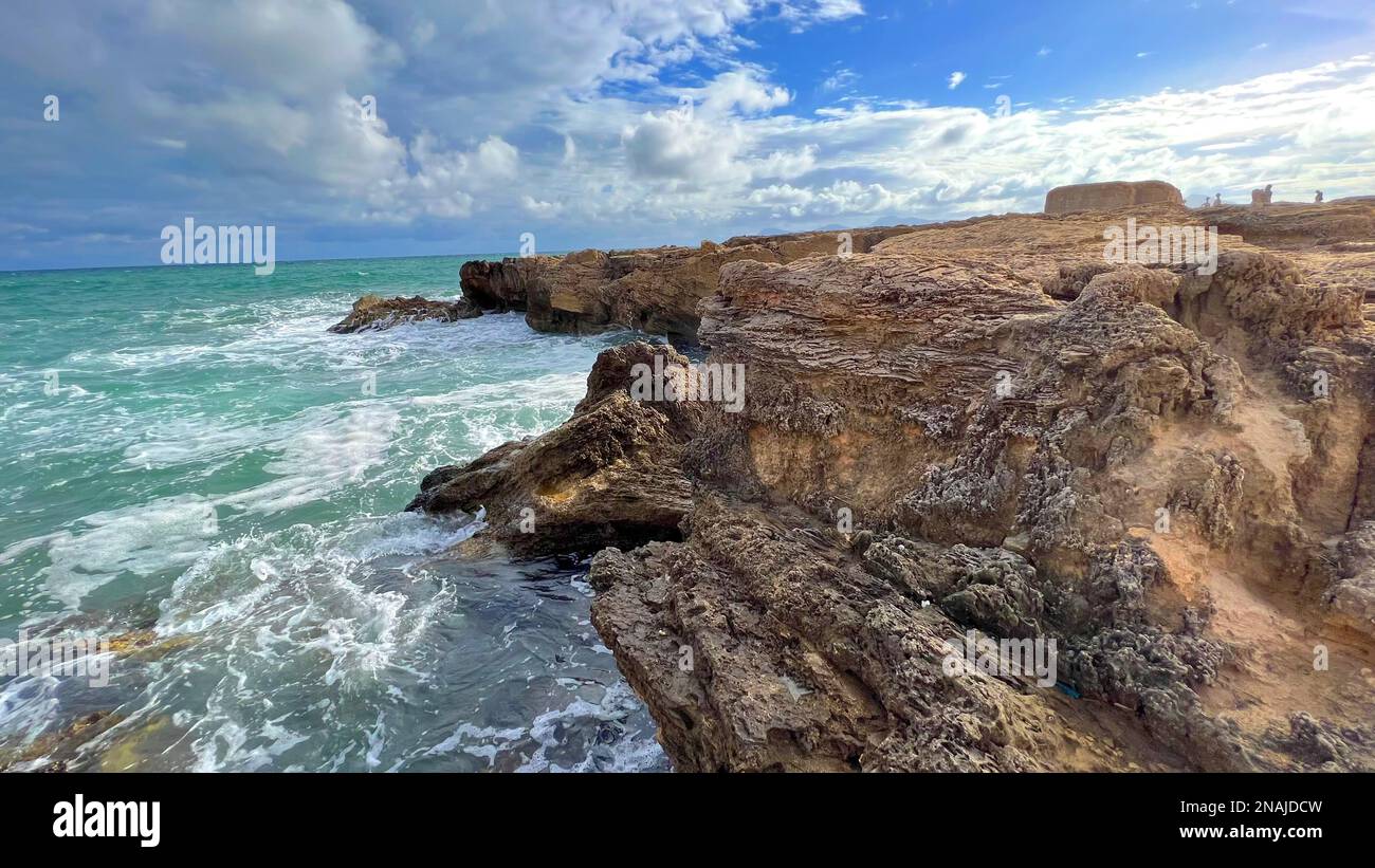 The sea waves crash into the rocky coast under a blue sky with clouds ...