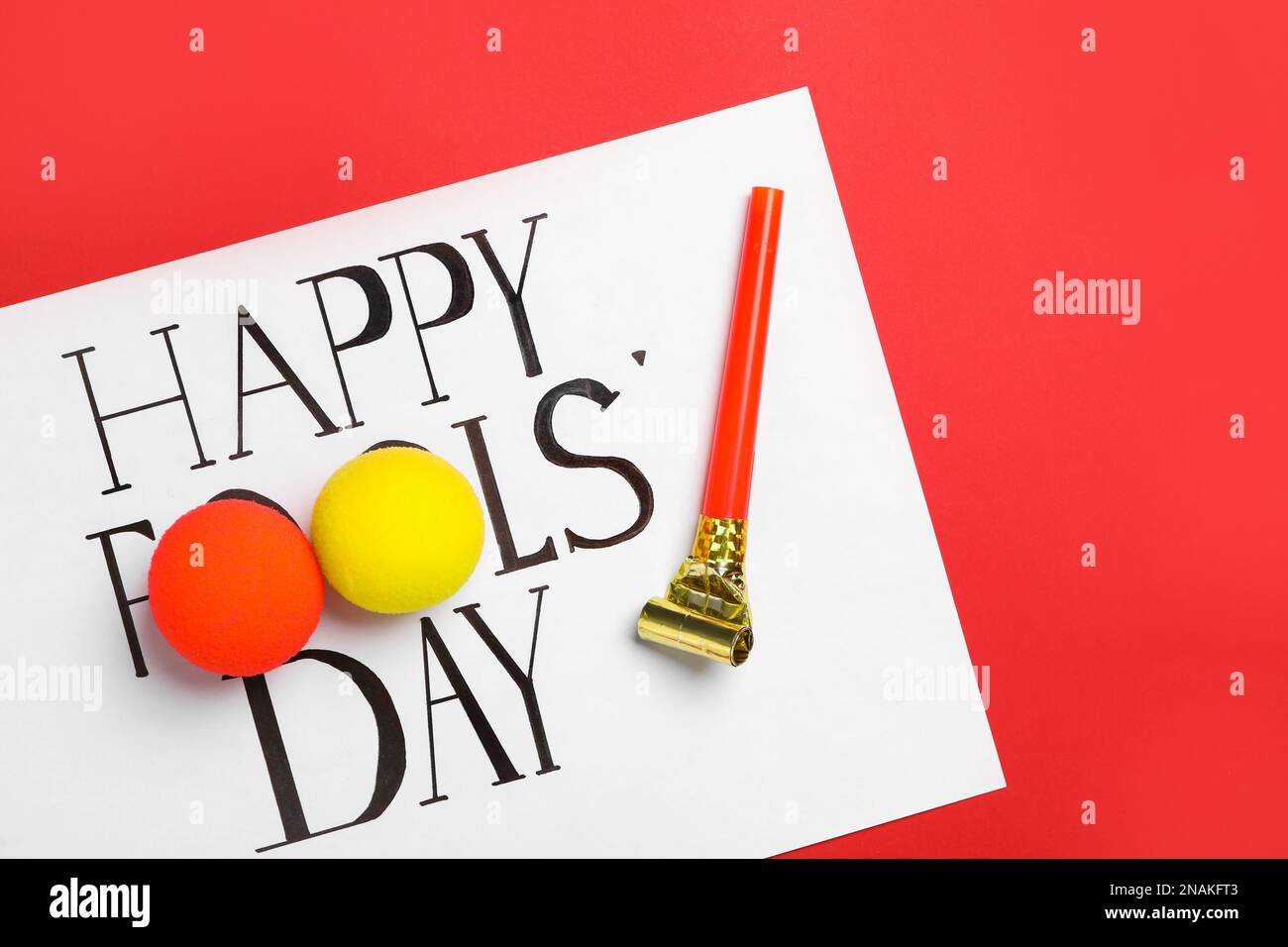 Sheet of paper with phrase Happy Fools' Day, clown noses and party blower on red background, flat lay Stock Photo
