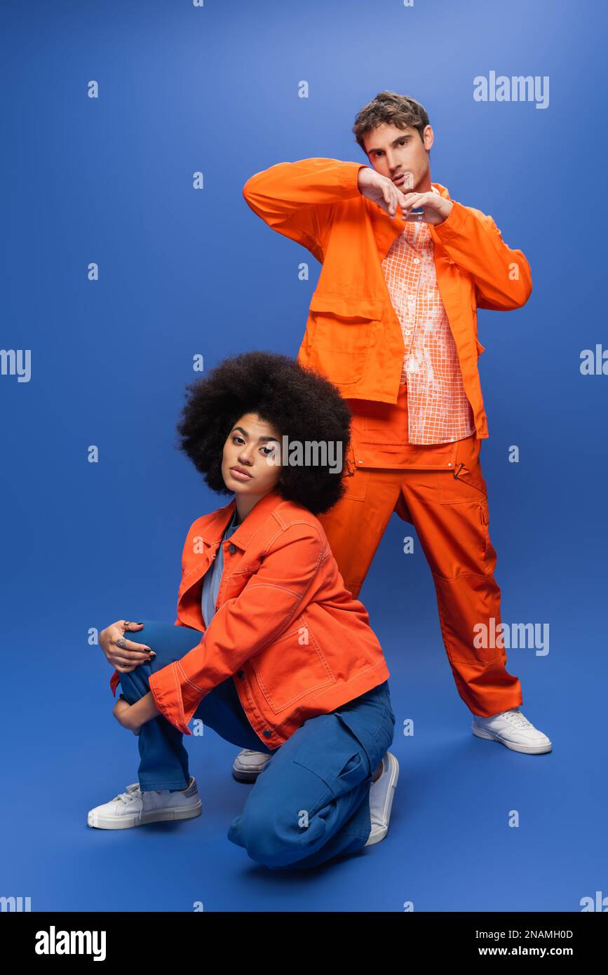 Trendy man in overalls posing near african american woman in orange jacket on blue background,stock image Stock Photo