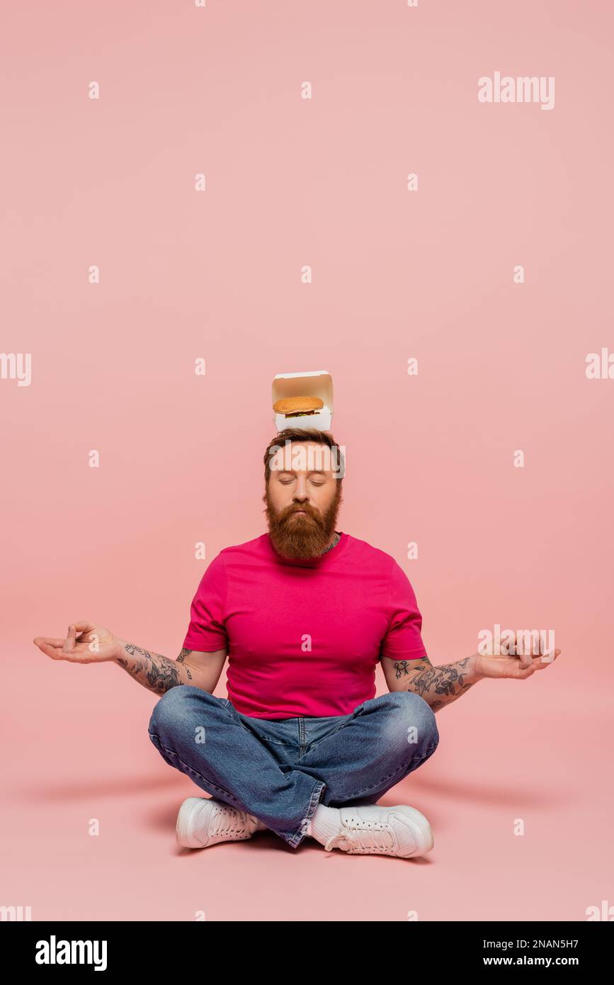 stylish bearded man with closed eyes and carton box with tasty burger on head meditating in lotus pose on pink background,stock image Stock Photo