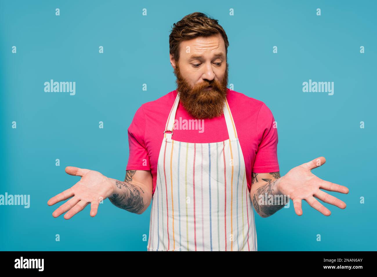 upset bearded man in striped apron looking at empty hands isolated on blue,stock image Stock Photo