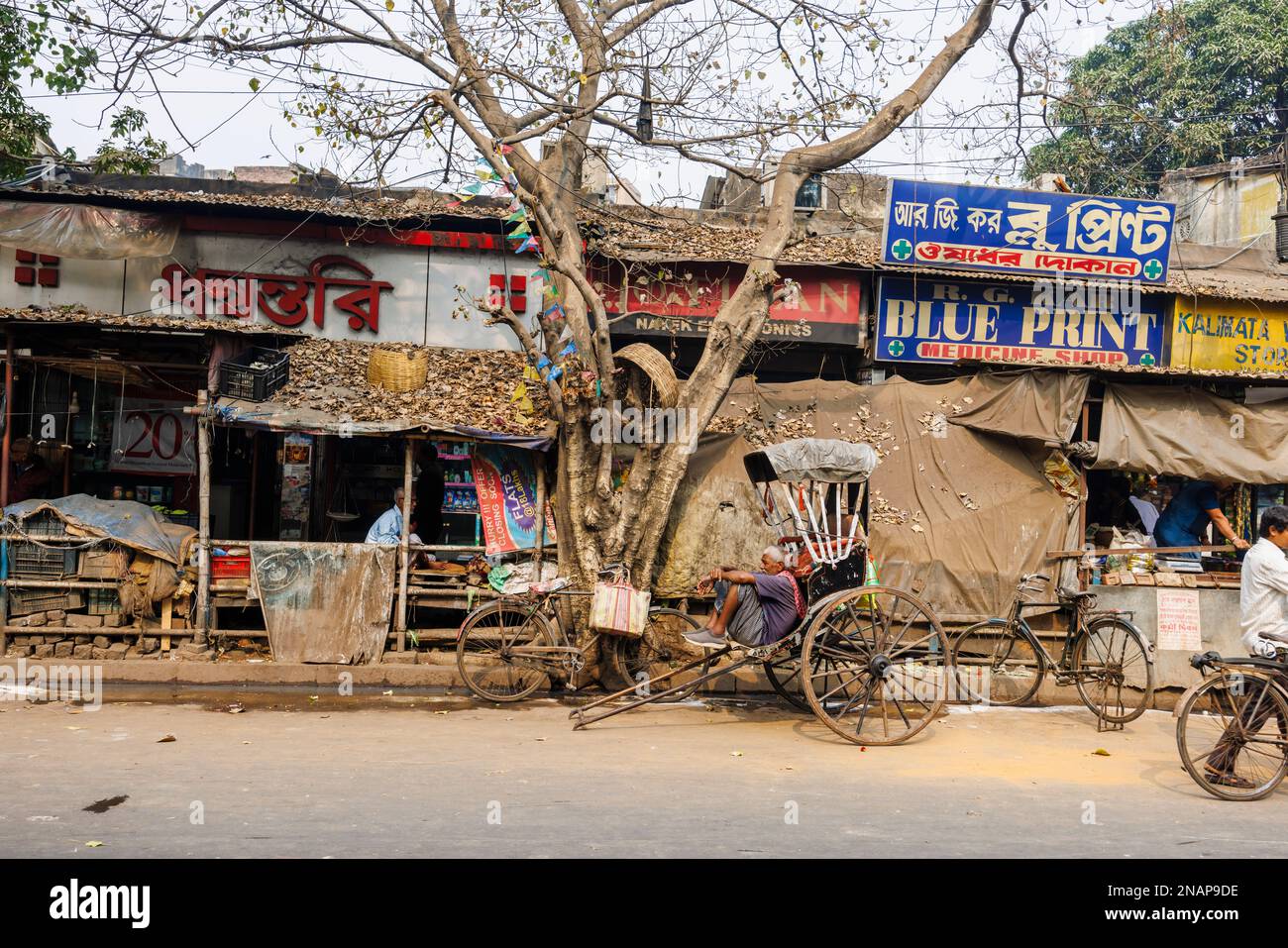 Street scene of a resting rickshaw driver, local people, shops, stalls and kiosks in Fariapukur, Shyam Bazar, a suburb of Kolkata, West Bengal, India Stock Photo