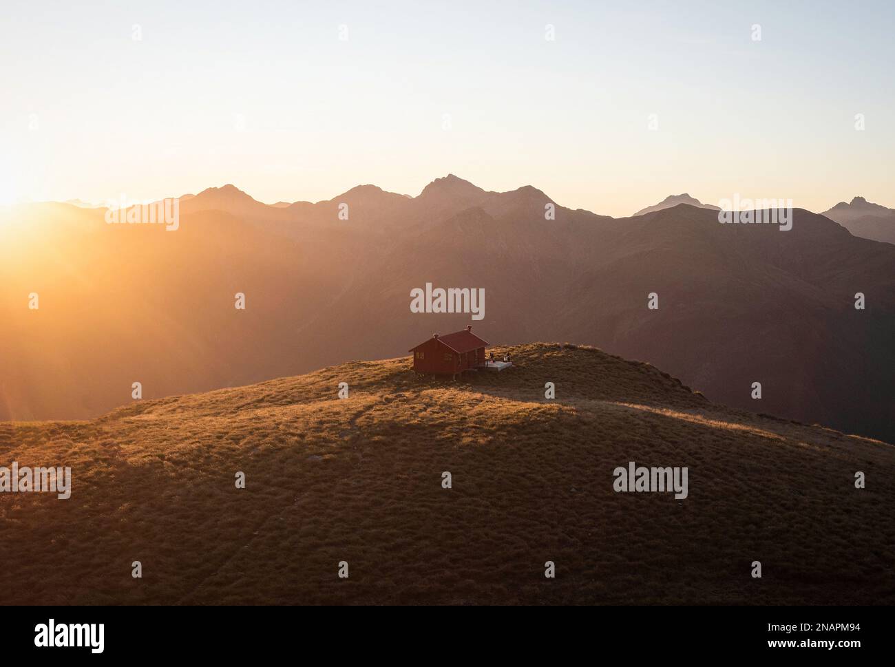 Sunset panorama of red idyllic remote lonely alpine mountain Brewster Hut perched on top of grass hill high up above Haast river valley, West Coast So Stock Photo