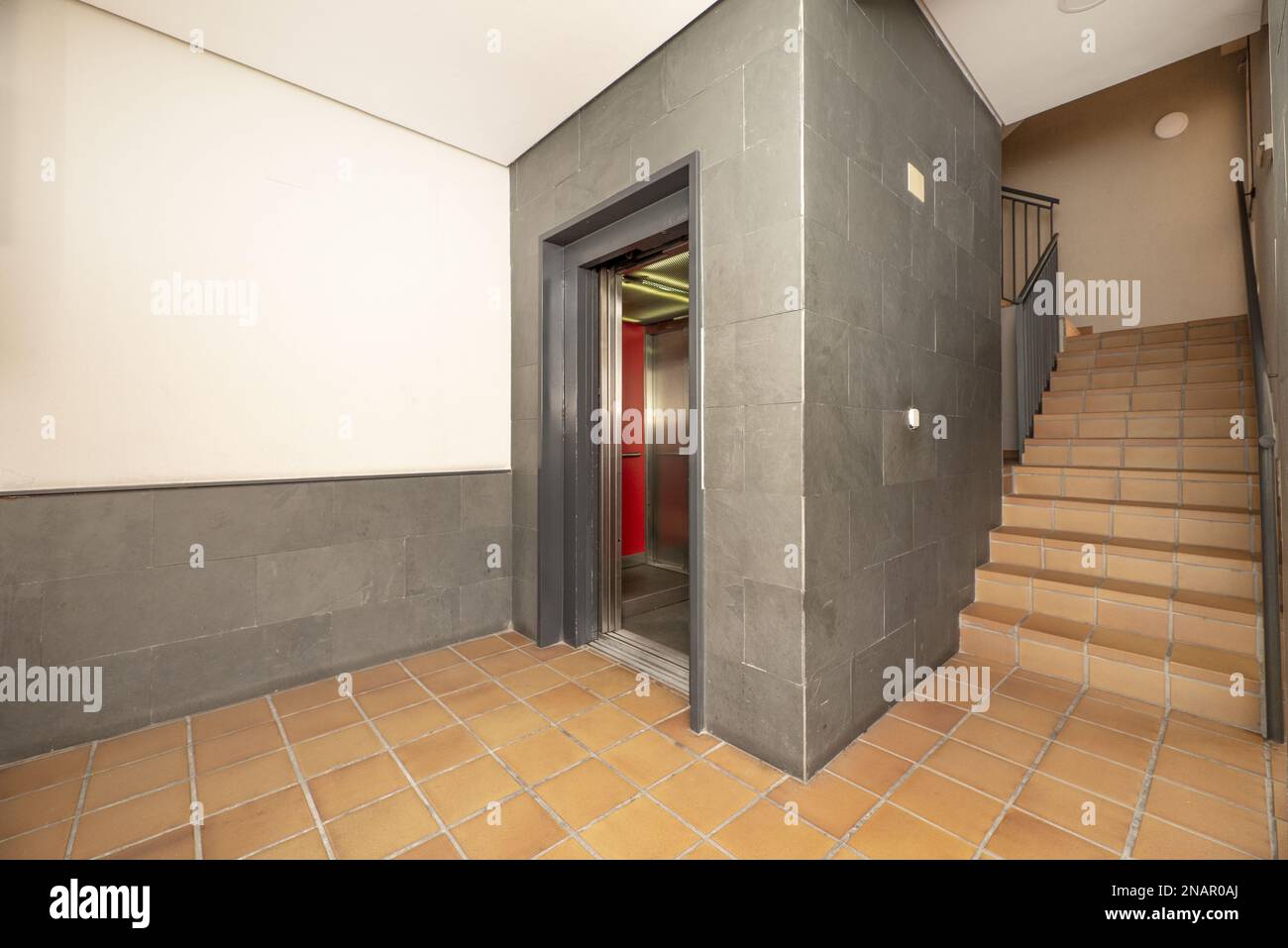 Interior of a residential housing portal with an elevator with open metal doors, stairs and a brown stoneware floor Stock Photo