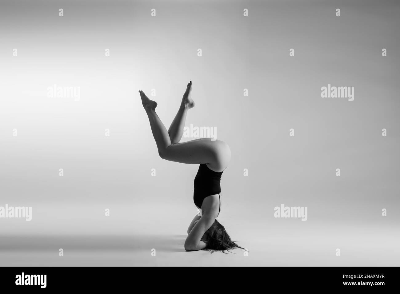 Young slim female in black bodysuit makes yoga stretching exercise on white studio wall background. Stock Photo