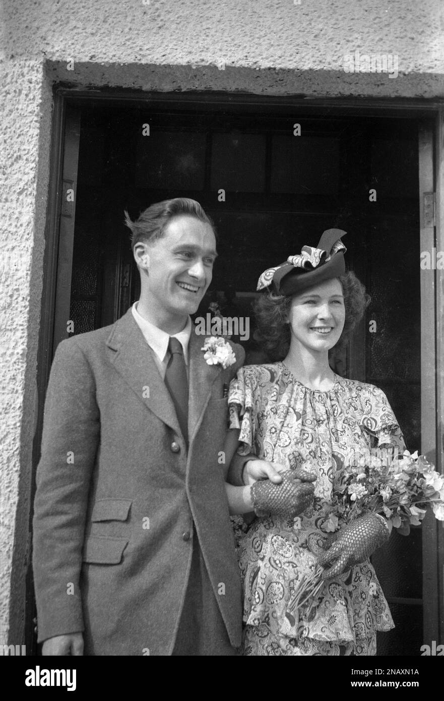 1950s, historical, a recently married couple, standing together in a doorway, smiling, happy and holding hands, the woman wearing lace gloves, floral dress and pretty hat, England, UK. Stock Photo