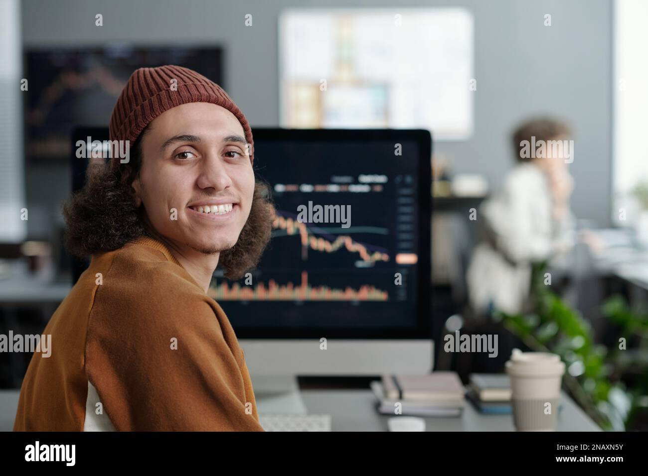 Happy young IT support manager looking at camera while sitting by workplace in front of computer screen with graphic data Stock Photo
