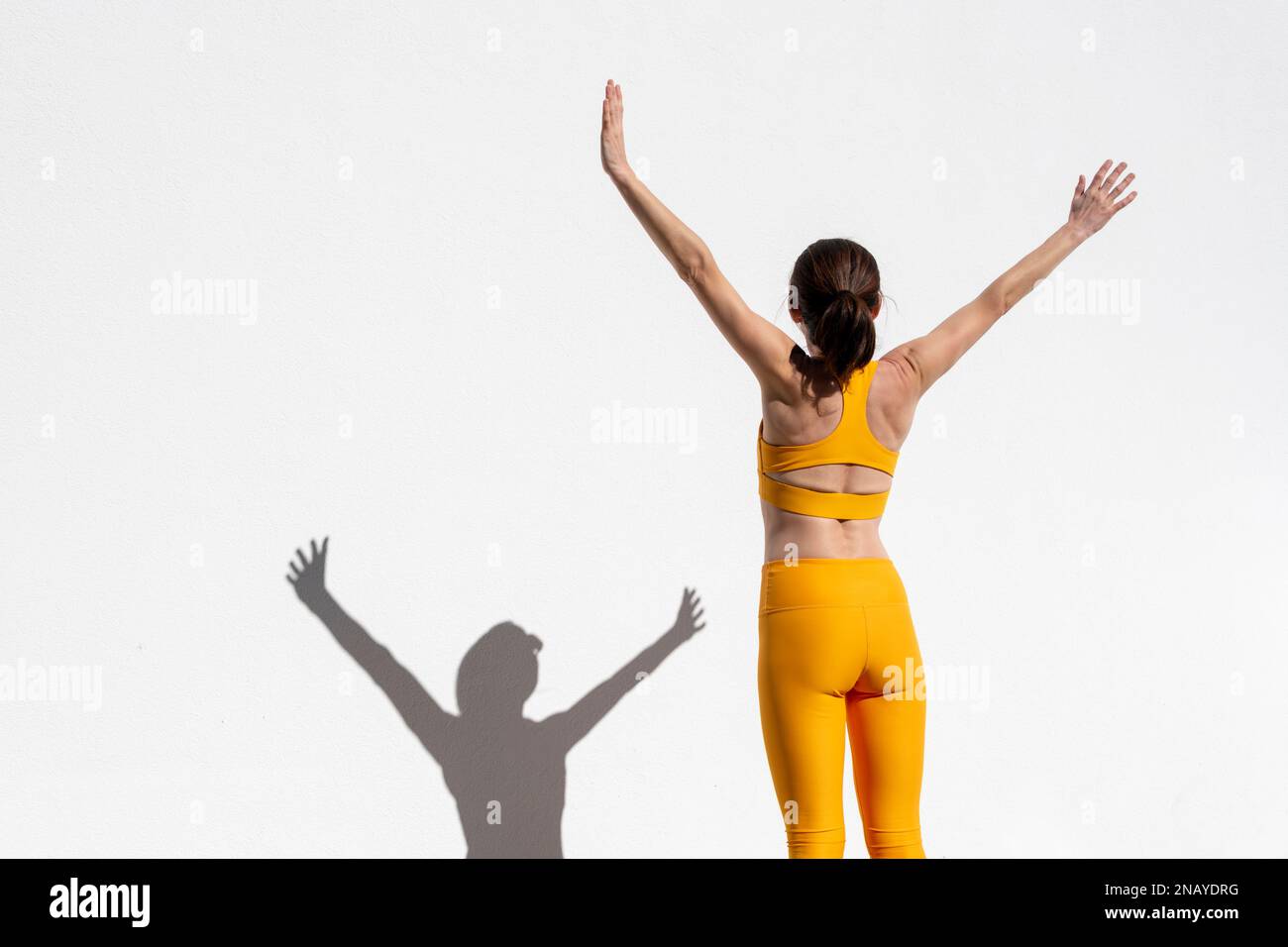 Rear view of a fit, sporty woman with her arms raised standing by a white wall, fitness concept. Stock Photo