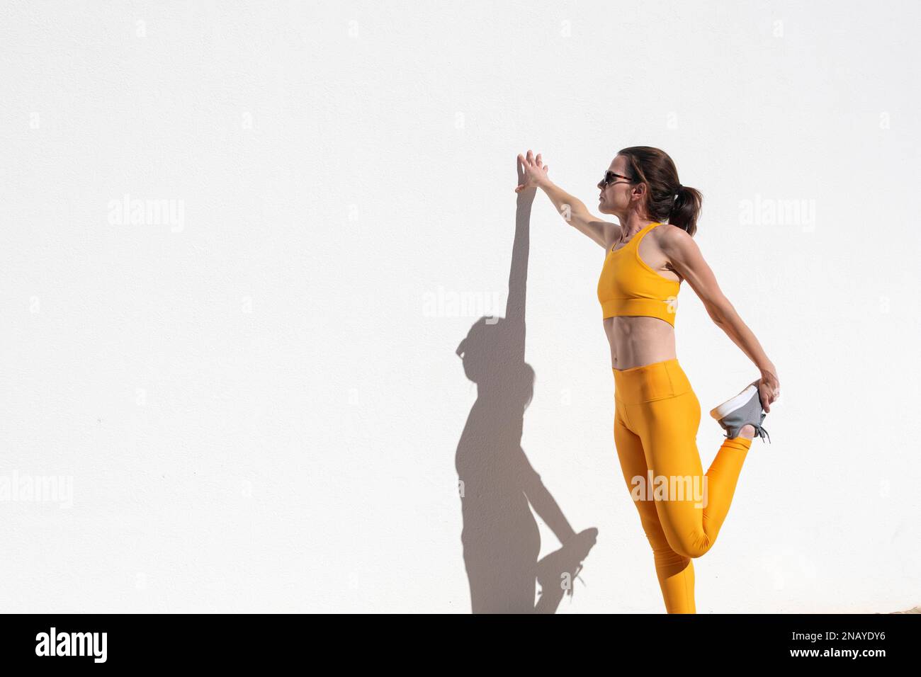 Sporty woman doing a leg stretch warm up exercise against a white wall. Stock Photo