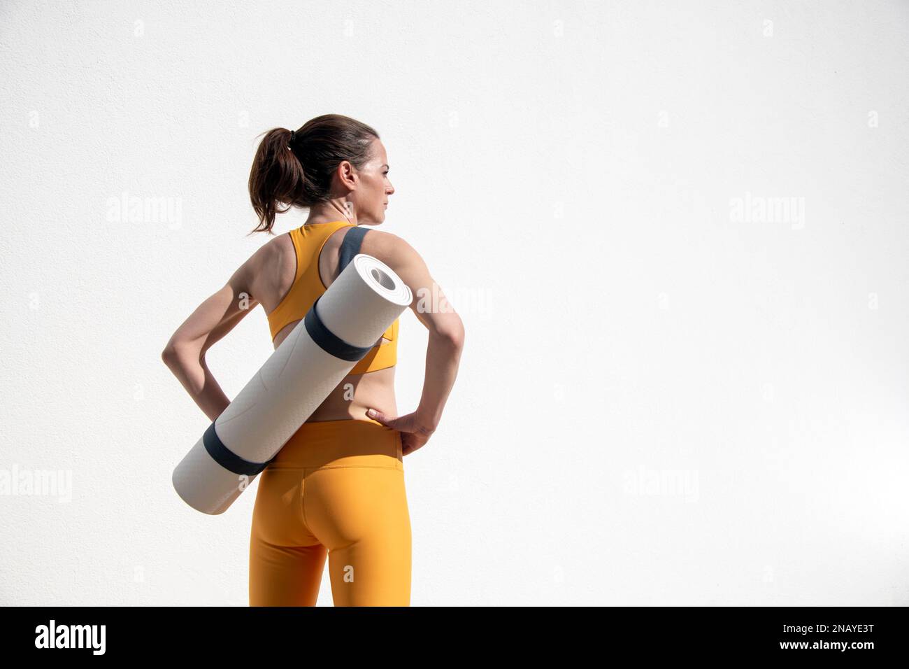 Sporty female carrying a yoga, exercise mat, outside by a white wall. Stock Photo