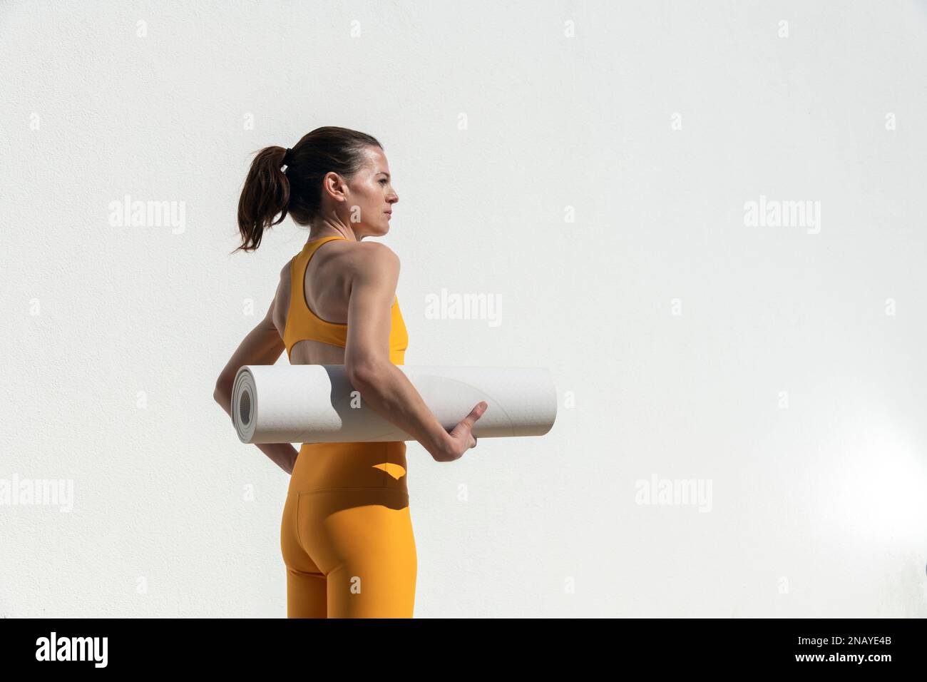 Sporty female carrying a yoga, exercise mat, outside by a white wall. Stock Photo