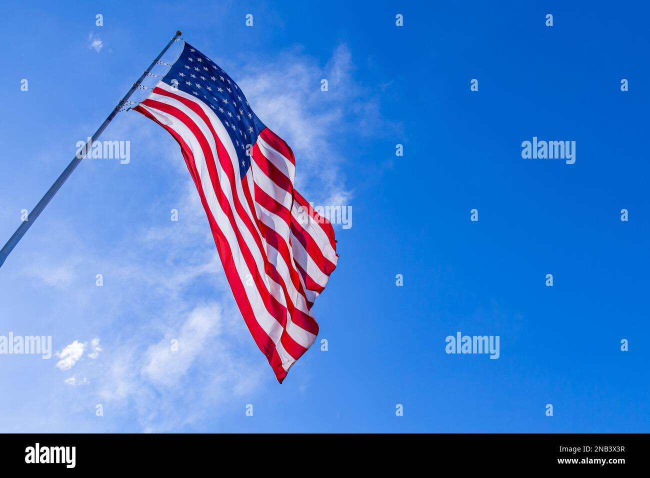 Close up of large American flag waving in front of blue sky and white cloud. American Flag waving in wind. Close up of United States flag. American Flag Waving Under A Blue Sky With Clouds. Stock Photo