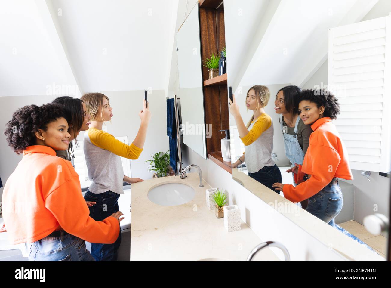 Happy diverse teenage female friends taking selfie in front of mirror. Spending quality time at home concept. Stock Photo