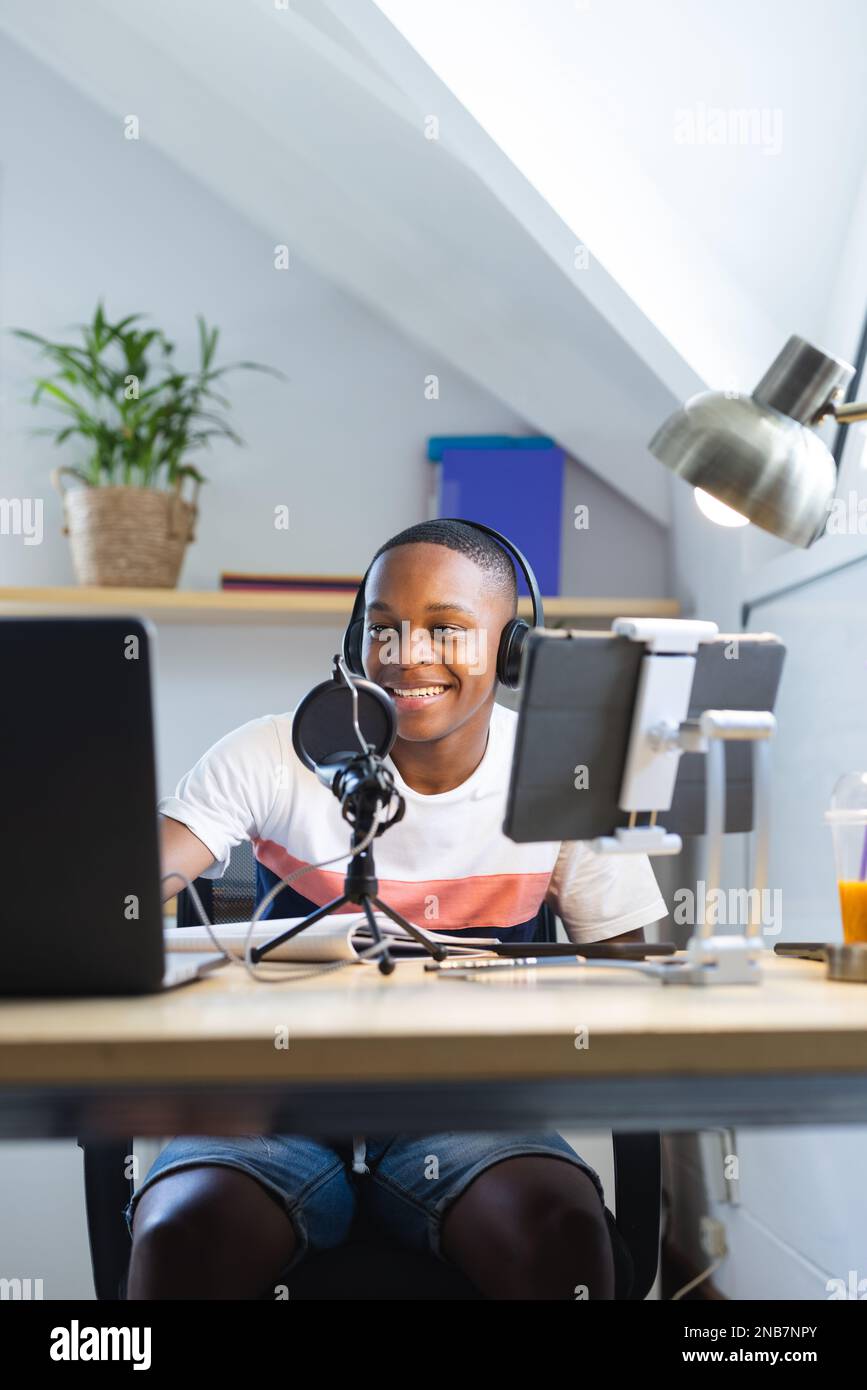 Vertical image of happy african american teenager with headphones and microphone recording podcast. Teenager, adolescence, hobby and spending free tim Stock Photo