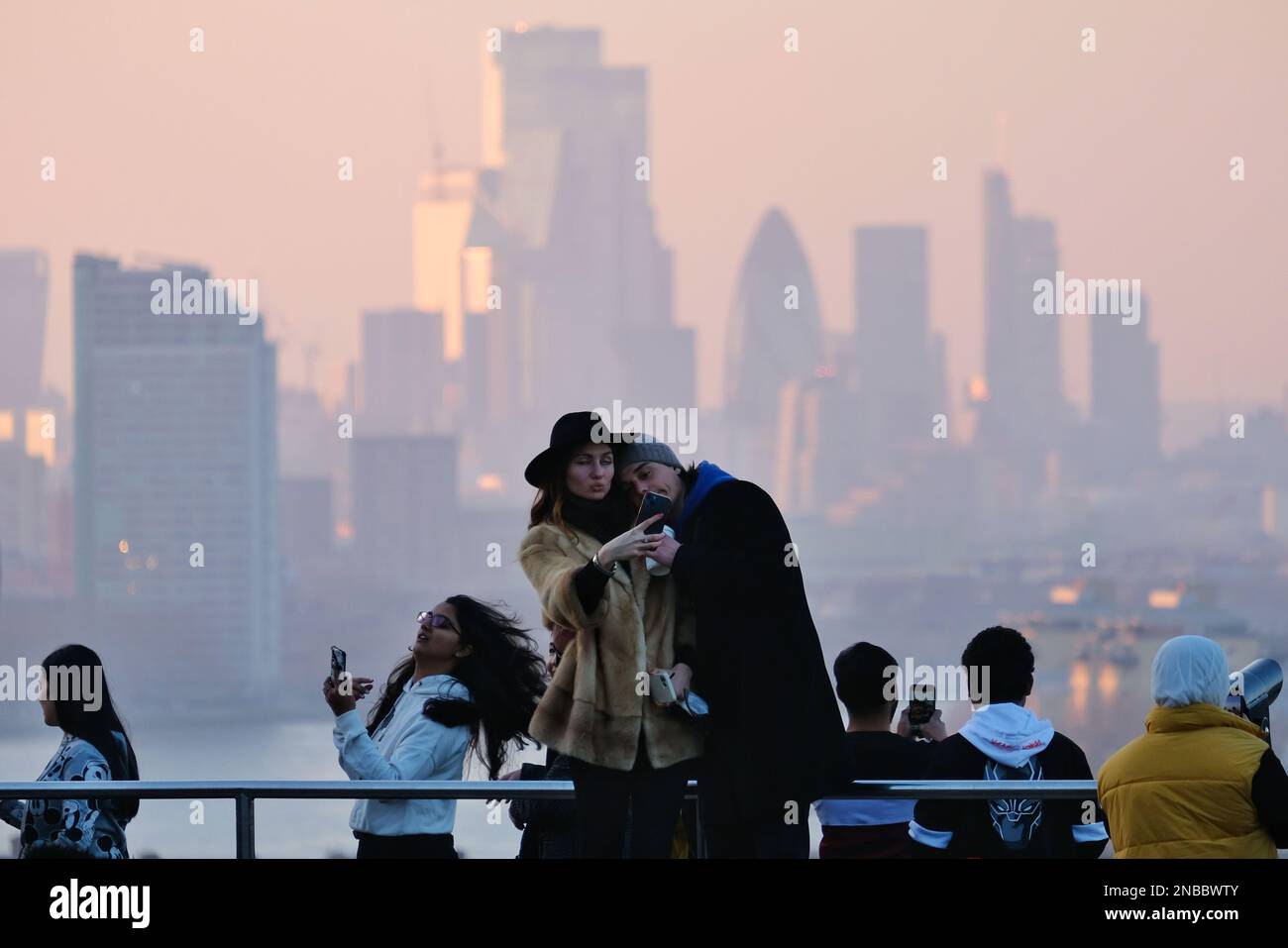 London, UK. 13th February, 2023. A couple take a selfie with the City of London buildings as a backdrop. Hazy conditions were observed at Greenwich Park during sunset and dusk, as southerly winds are expected to bring Saharan dust deposits to the UK, which will peak between Tuesday and Wednesday. The mineral dust in the atmosphere can turn skies an orange hue, cause 'blood rain' and spectacular sunrises and sunsets. Credit: Eleventh Hour Photography/Alamy Live News Stock Photo