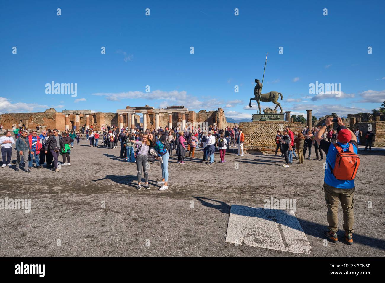 Groups of tourists, visitors pack, crowd the forum area, busy posing for photographs, selfies. At Pompeii Archaeological Park near Naples, Italy. Stock Photo